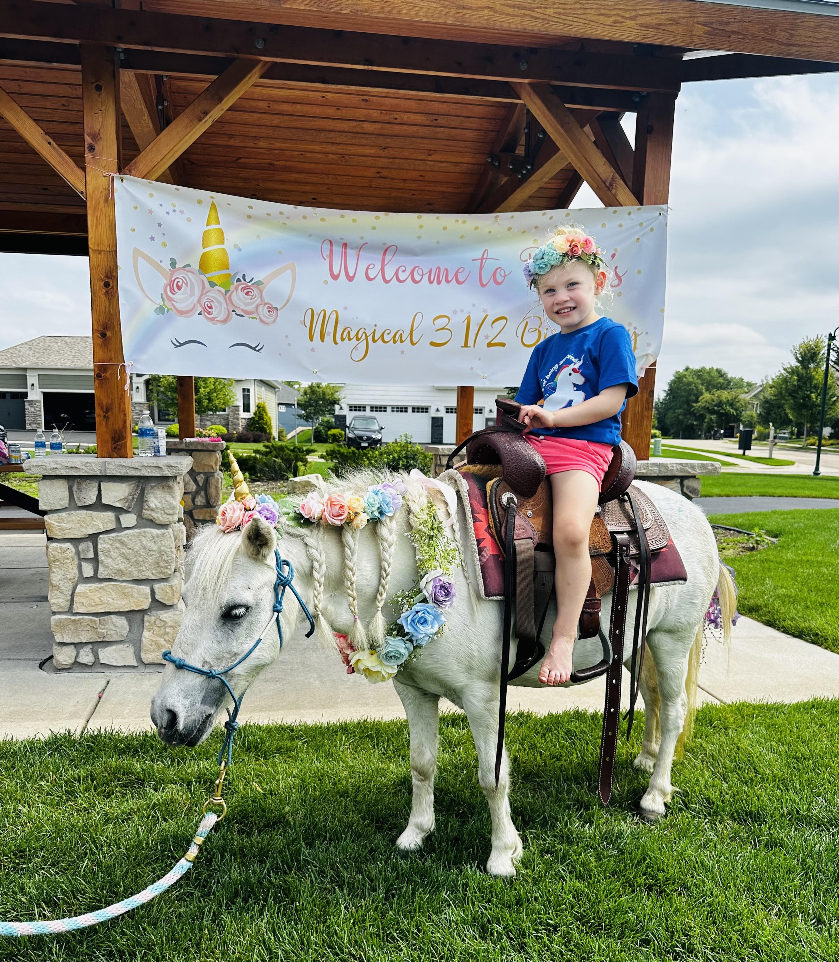 A young girl in a flower crown sitting in the saddle on a white pony decorated with braided, flower-woven mane at a backyard birthday party